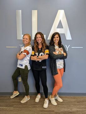Three LLA Therapy team members posing in football jerseys in front of the LLA logo on a wall