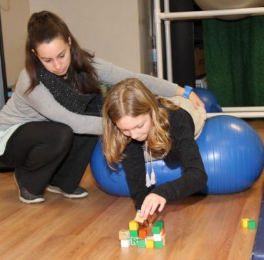 Therapist helping a girl who is lying over a blue therapy ball while she stacks wooden blocks