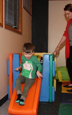 Boy sliding down a colorful indoor slide while a therapist stands nearby