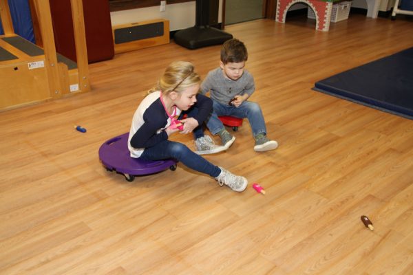 Two children playing on scooter boards on a wood floor in a therapy gym