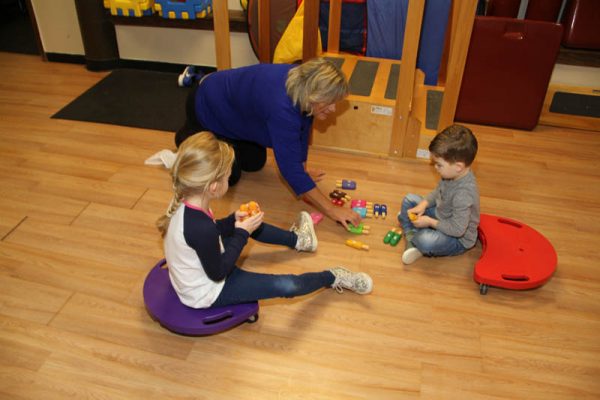 Occupational therapist kneeling on the floor playing with building toys with two children on scooter boards