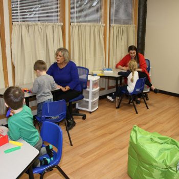 Two therapists working with small groups of children at tables in a bright therapy room with a green bean bag chair