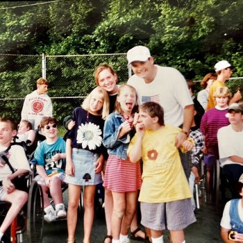 Group of children and teens, some using wheelchairs, posing with adults outdoors at a camp
