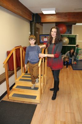 Boy walking down a therapy staircase with a handrail while a therapist stands beside him in a therapy gym