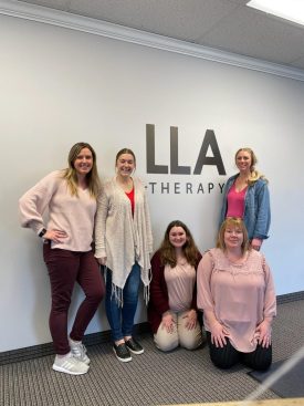Five LLA Therapy team members posing in front of the LLA Therapy logo on a wall