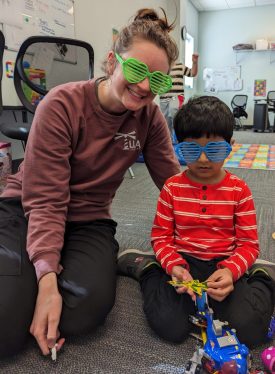 Therapist and a boy in a red striped shirt, both wearing sunglasses, playing with toys on the floor
