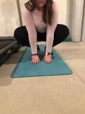 Person in a deep squat with hands flat on a teal mat, demonstrating a frog jump