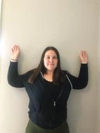 Woman standing against a wall with both arms raised and bent in a W shape to demonstrate the W reach stretch
