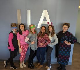 Six LLA Therapy team members posing in front of the LLA logo on a wall, some wearing silly sleep masks and glasses